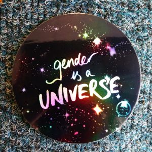 Rainbow holographic stickers showing "gender is a UNIVERSE" design on it in holographic plastic